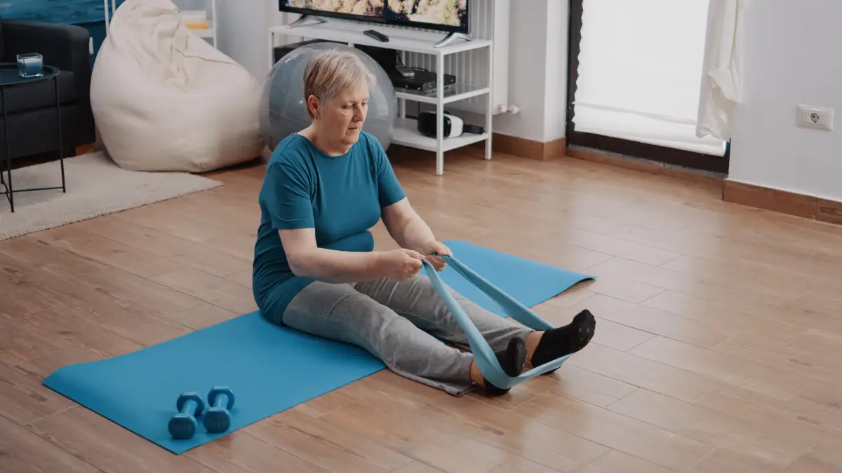 Eine ältere Frau sitzt auf einer Trainingsmatte in einem Wohnzimmer und führt mit einem Widerstandsband eine Rehabilitationsübung für die Beine durch. Neben ihr liegen Hanteln auf dem Boden, und im Hintergrund sind ein Fernseher sowie ein Gymnastikball zu sehen.