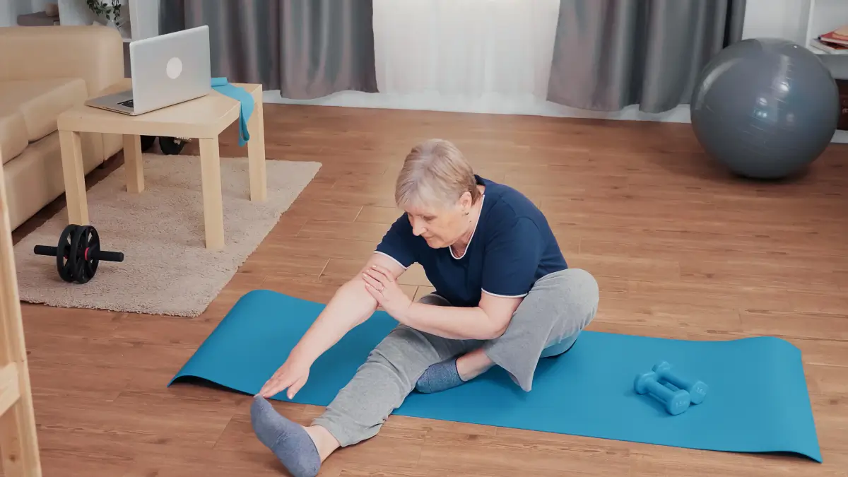 Das Bild zeigt eine ältere Frau, die auf einer blauen Yoga-Matte sitzt und sich nach vorne beugt, um ihr Bein zu dehnen. Sie befindet sich in einem gemütlich eingerichteten Raum mit Holzboden. Im Hintergrund sind Fitnessgeräte wie eine Hantel, ein Gymnastikball und ein Laptop auf einem Tisch zu sehen, was auf ein Heimtraining hindeutet. Die Szene vermittelt eine aktive und gesunde Lebensweise im Alter.