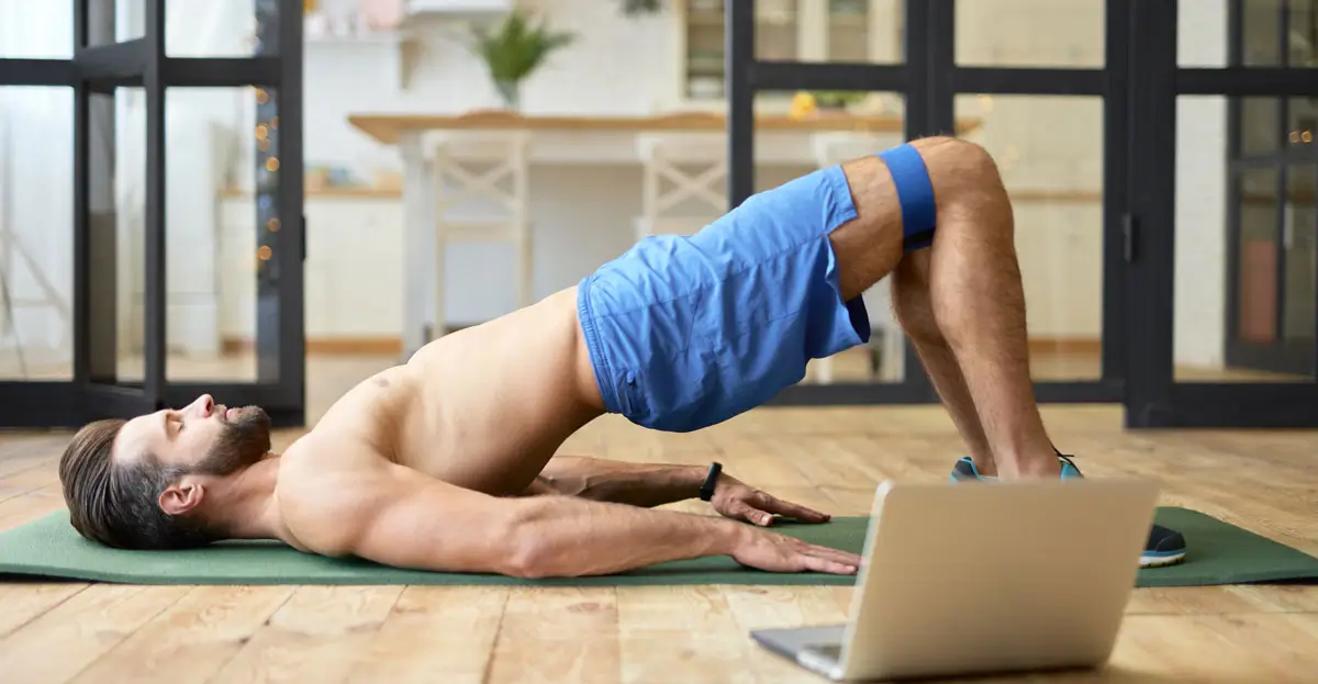 Ein athletischer Mann mit nacktem Oberkörper führt eine Brückenübung auf einer Yogamatte in einem gemütlichen Raum durch. Er verwendet ein Widerstandsband um die Oberschenkel und hat einen Laptop vor sich, was darauf hindeutet, dass er einem virtuellen Workout-Programm folgt. Die Szene vermittelt Fitness und Heimtraining.