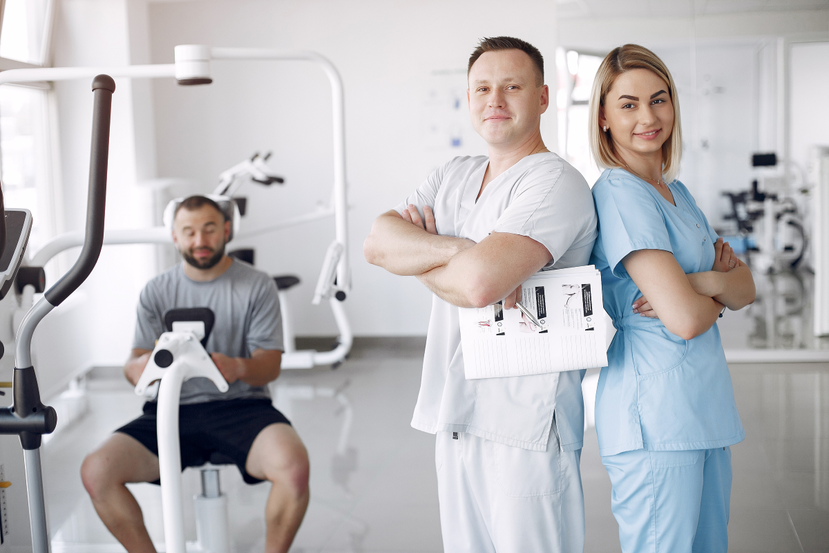 Das Bild zeigt zwei medizinische Fachkräfte, einen Mann und eine Frau, die selbstbewusst mit verschränkten Armen in einer physiotherapeutischen Klinik stehen. Der Mann hält einen Klemmbrett mit Dokumenten. Im Hintergrund sieht man einen Patienten, der auf einem Fitnessgerät sitzt, sowie weitere Trainings- und Rehabilitationsgeräte. Die Szene vermittelt den Eindruck von Professionalität und Unterstützung in einem gesundheitlichen Umfeld, das auf Rehabilitation und Fitness ausgerichtet ist.