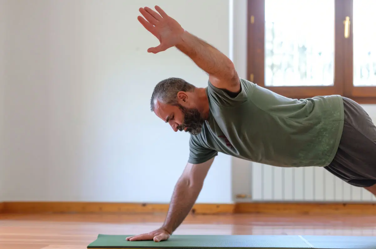 Ein sportlicher Mann führt eine Yogaübung in einem hellen Raum durch. Er stützt sich auf einer Yogamatte ab, während er einen Arm ausgestreckt hebt. Die Szene zeigt Konzentration, Körperbeherrschung und eine ruhige, minimalistische Umgebung, die zur Achtsamkeit einlädt.