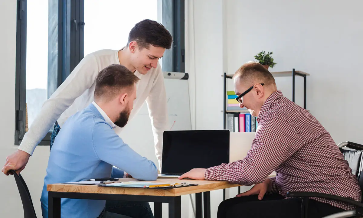 Drei Männer arbeiten gemeinsam in einem modernen Büro an einem Tisch. Sie sind in eine Diskussion vertieft und betrachten konzentriert den Bildschirm eines Laptops. Die Szene vermittelt Teamarbeit, Zusammenarbeit und eine produktive Atmosphäre.