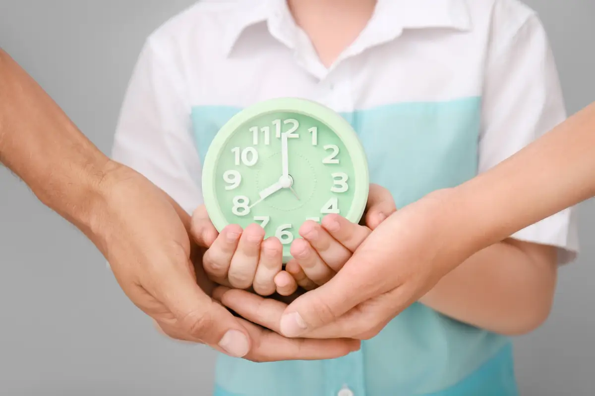 Hände von Erwachsenen und einem Kind halten gemeinsam eine grüne Uhr. Die Szene symbolisiert Zusammenarbeit, Zeitmanagement oder die Bedeutung von gemeinsam verbrachter Zeit. Im Hintergrund ist ein Kind in einem weißen Hemd mit türkisen Details zu sehen.