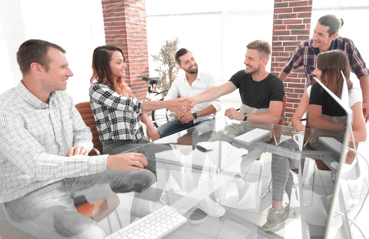 Gruppe junger Kolleginnen und Kollegen in einem modernen Büro, die an einem Glastisch sitzen. Zwei Personen in der Mitte geben sich die Hand, während die anderen lächelnd zusehen. Die entspannte Atmosphäre deutet auf Teamarbeit, Zusammenarbeit oder einen erfolgreichen Abschluss hin.