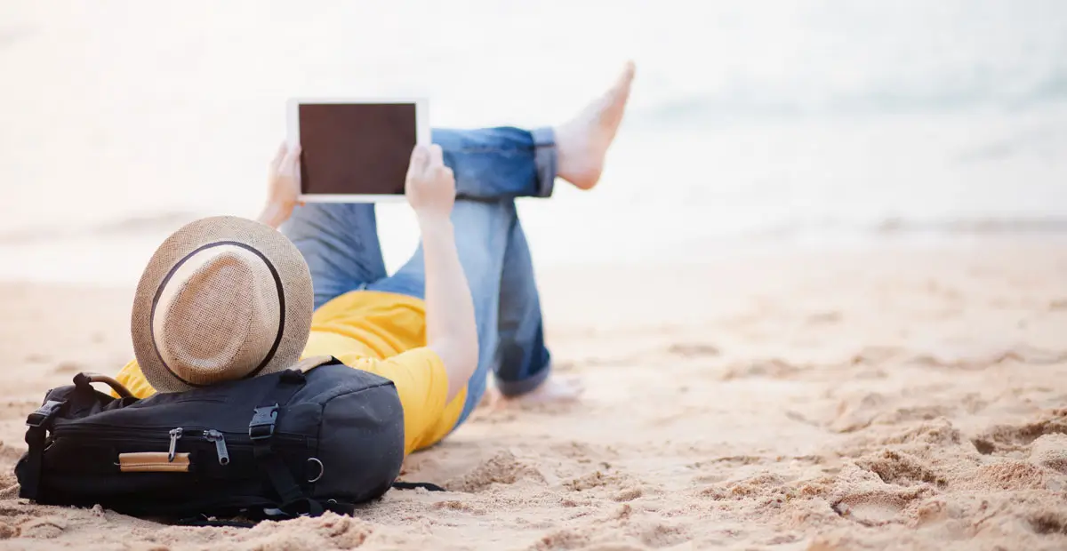 Eine Person liegt entspannt am Strand, trägt einen Strohhut und hält ein Tablet in der Hand. Neben ihr liegt ein schwarzer Rucksack. Im Hintergrund ist das Meer zu sehen, das eine ruhige und entspannte Atmosphäre vermittelt.