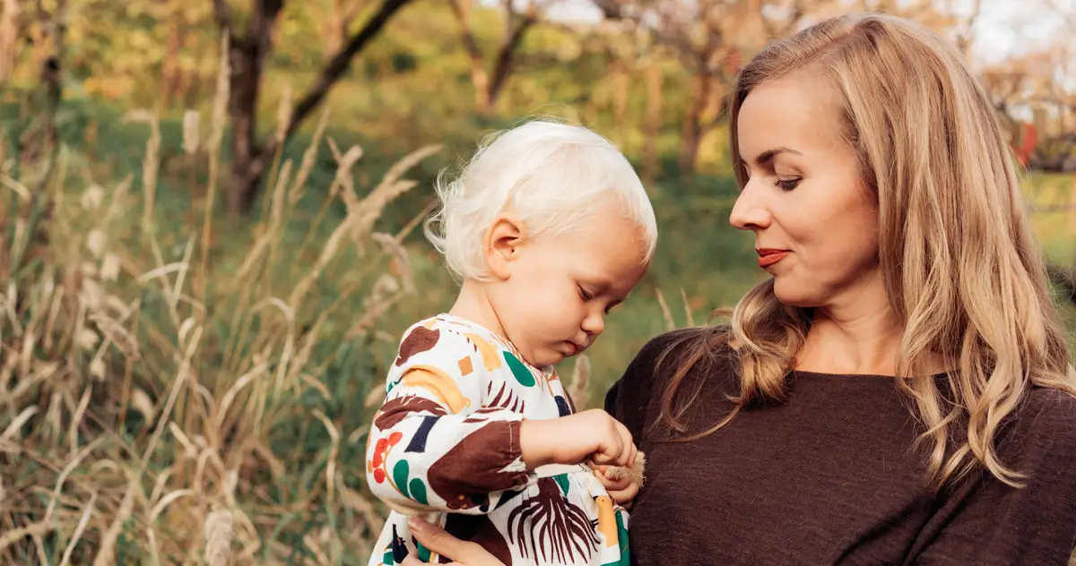 Eine Mutter hält liebevoll ihr kleines Kind mit blondem Haar im Arm, während sie in einem natürlichen, grasbewachsenen Feld stehen. Das Kind trägt ein farbenfrohes Kleid und schaut konzentriert auf etwas in seiner Hand. Die Szene vermittelt Geborgenheit, Naturverbundenheit und familiäre Wärme.