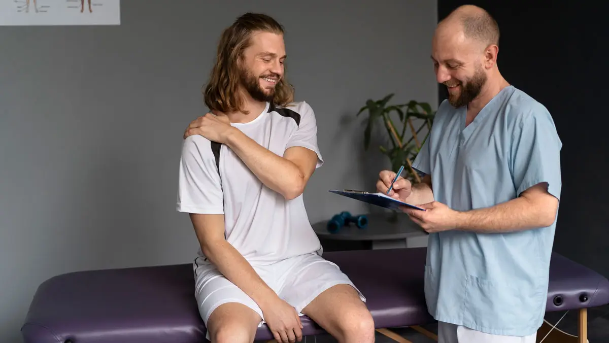 Ein Physiotherapeut in blauer Arbeitskleidung spricht lächelnd mit einem Patienten, der auf einer Behandlungsliege sitzt und seine Schulter hält. Beide wirken entspannt und freundlich. Im Hintergrund ist eine Pflanze und Trainingsausrüstung zu sehen, was auf eine professionelle und einladende Rehabilitationsumgebung hinweist.