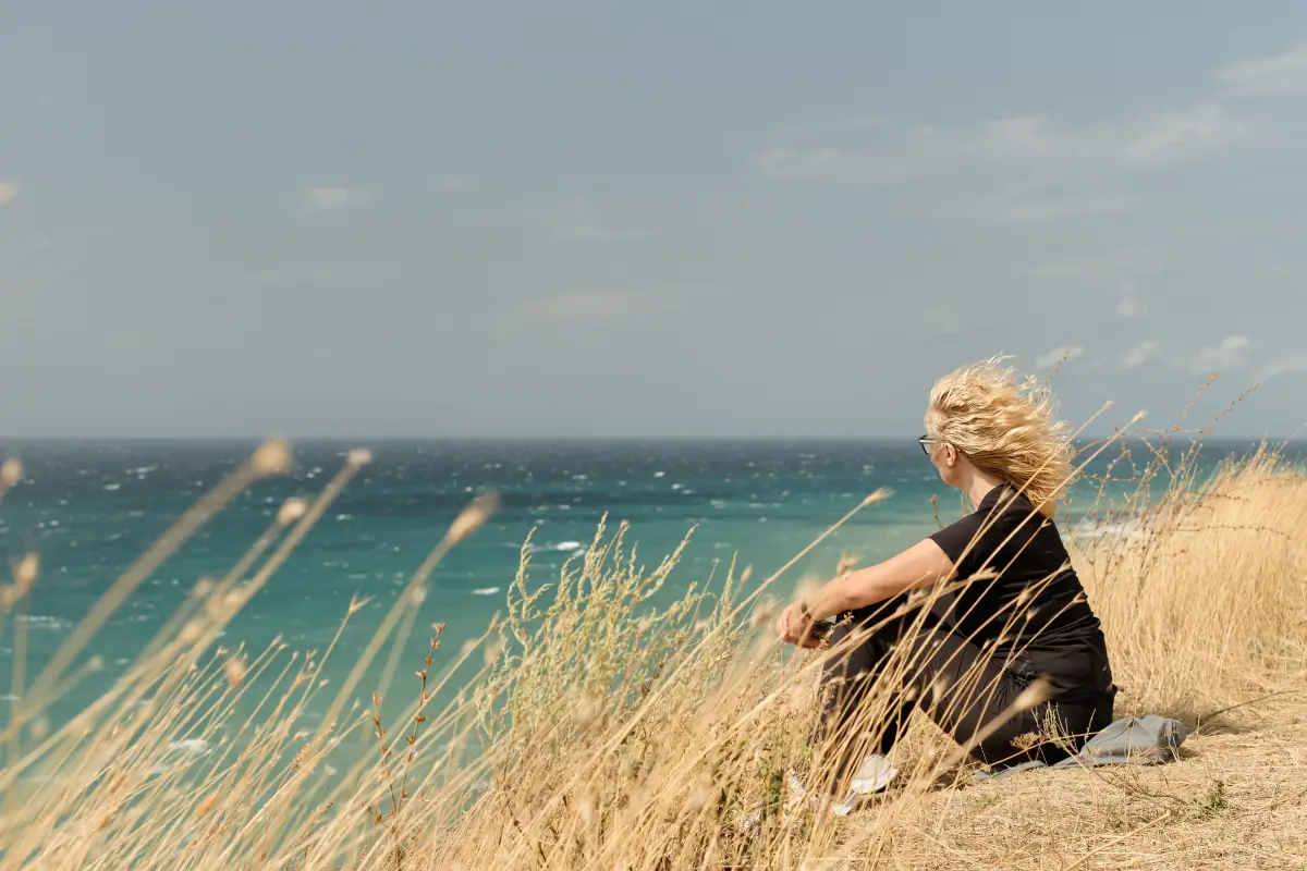 Eine Frau sitzt auf einem grasbewachsenen Hügel und blickt auf das weite, blaue Meer, während der Wind ihr Haar zerzaust.