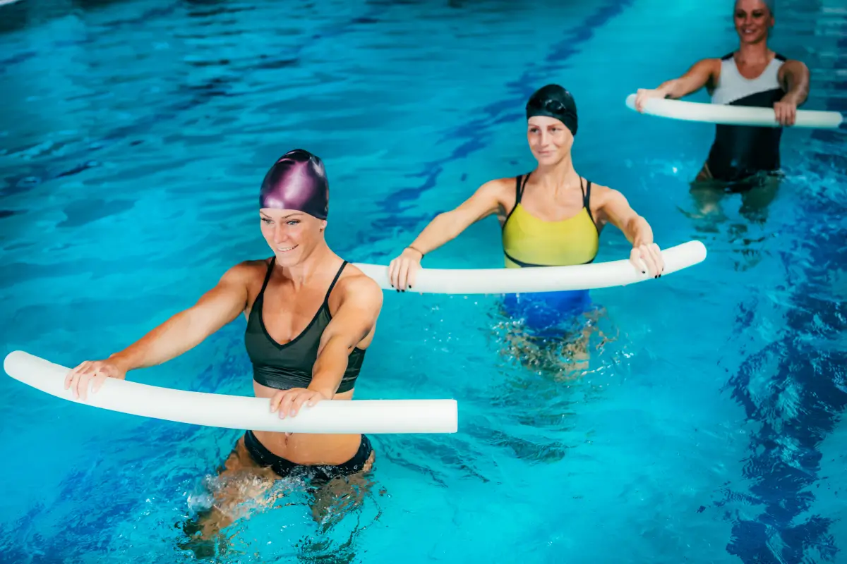 Frauen in einem Schwimmbad bei einer Aqua-Aerobic-Einheit, die synchron Übungen mit Schwimmnudeln durchführen.