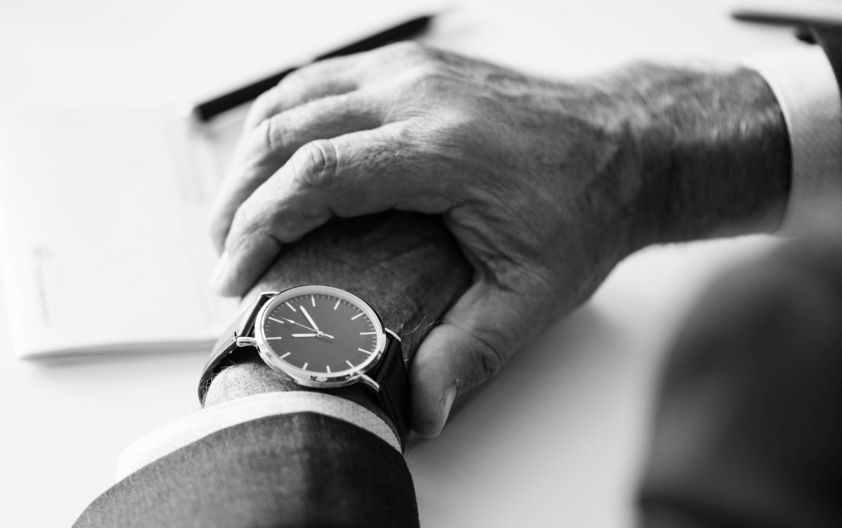 Eine Nahaufnahme einer älteren männlichen Hand, die eine andere Hand hält. Der Mann trägt eine elegante Armbanduhr, und im Hintergrund liegt ein Dokument auf dem Tisch.