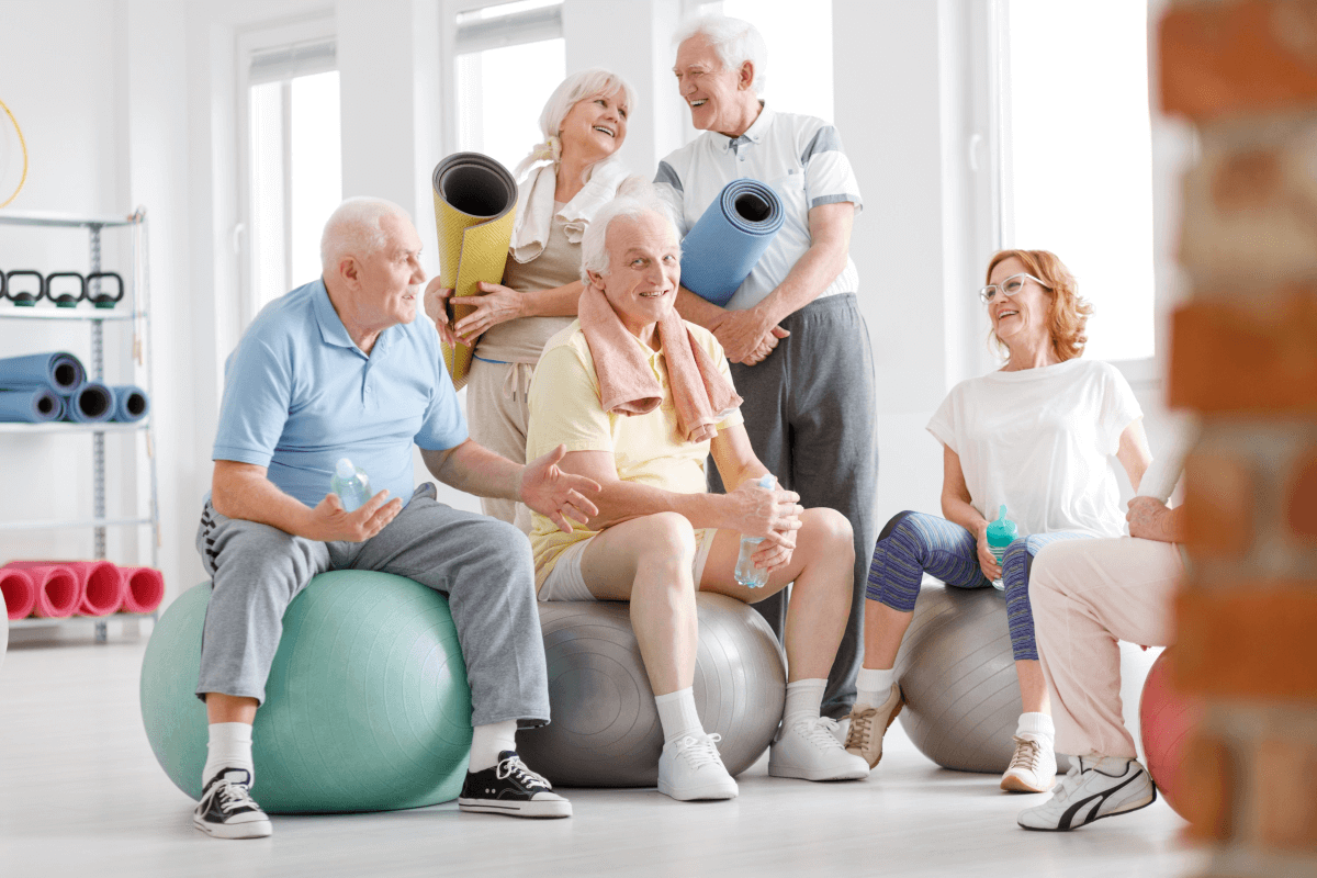 Eine Gruppe älterer Menschen sitzt auf Gymnastikbällen in einem hellen Fitnessstudio. Sie unterhalten sich fröhlich, während einige von ihnen Wasserflaschen halten oder Yogamatten tragen.