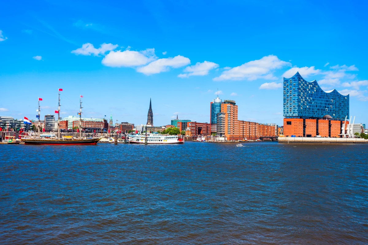 Panoramaansicht des Hamburger Hafens mit der Elbphilharmonie, historischen Segelschiffen und modernen Gebäuden vor blauem Himmel.