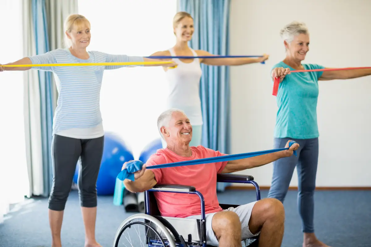 Eine Gruppe älterer Menschen macht Rehabilitationsübungen mit Therabändern, ein lachender Mann im Rollstuhl nimmt ebenfalls teil.