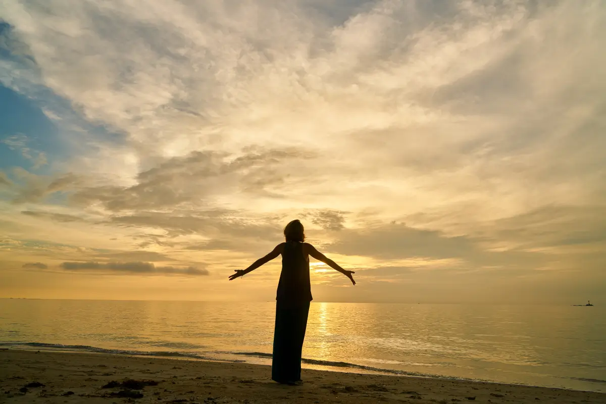 Silhouette einer Frau, die mit ausgestreckten Armen am Strand steht und den Sonnenuntergang über dem Meer genießt.