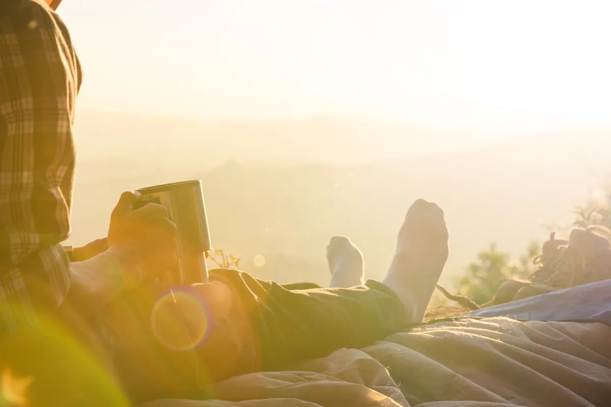 Junger Mann sitzt entspannt vor einem Zelt in der Morgensonne, hält eine Tasse Kaffee in der Hand und genießt den Blick auf eine Berglandschaft.